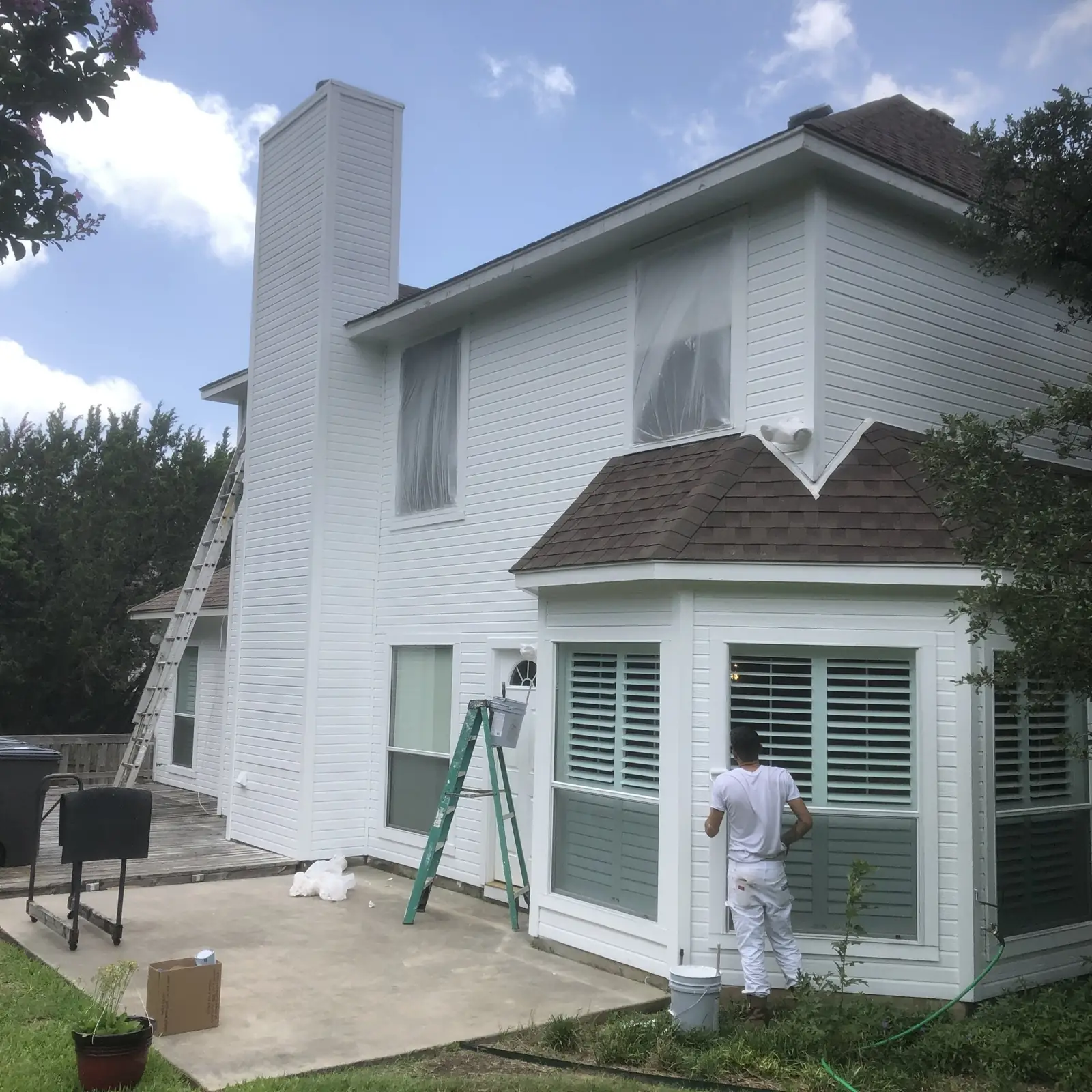 A painter on an extension ladder working on a white two story house with grey shutters, drop cloths spread on the ground