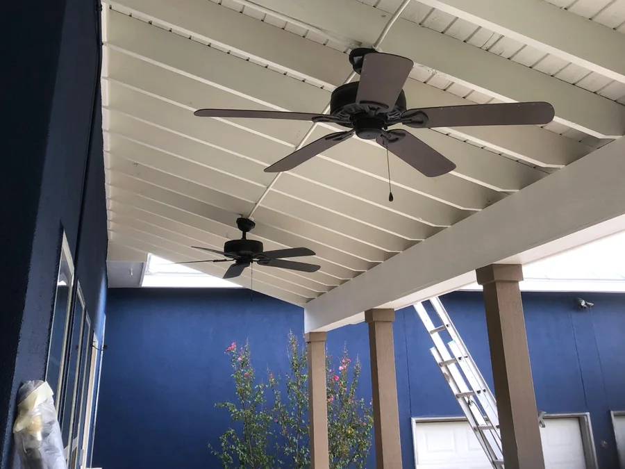 Covered porch mid project: white painted board ceiling and beams, tan columns, navy walls, masking on a window and a ladder still in place