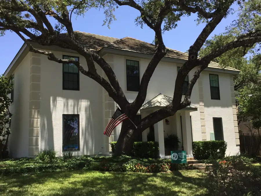 Two story light stucco home framed by a large live oak, with an American flag by the front door