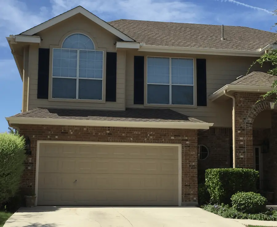 Two story home with tan siding, black shutters, brick and a double garage door under a clear sky