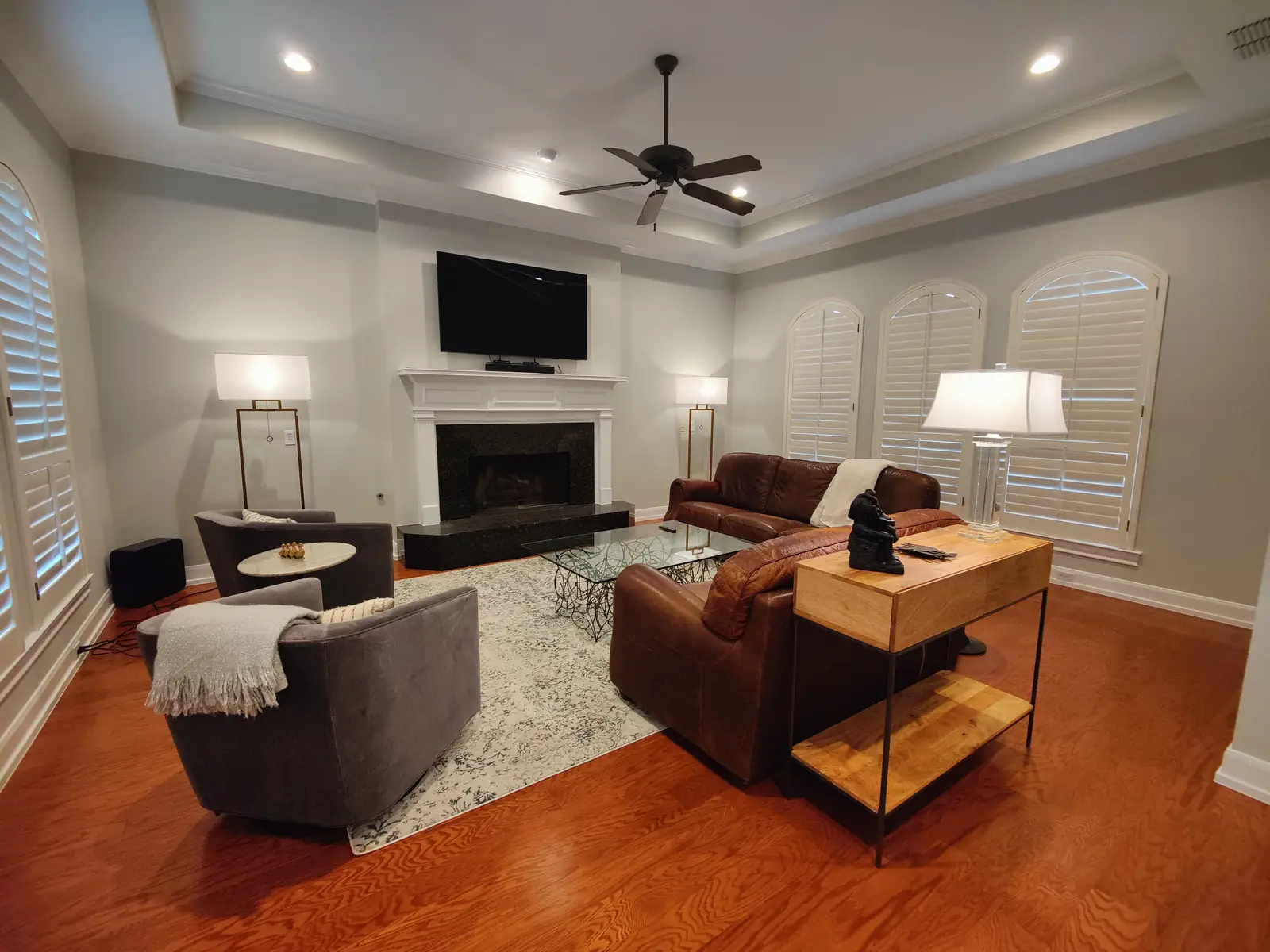 Living room with a tray ceiling, a white mantel with a black granite surround, arched plantation shutters, a brown leather sectional and a hardwood floor